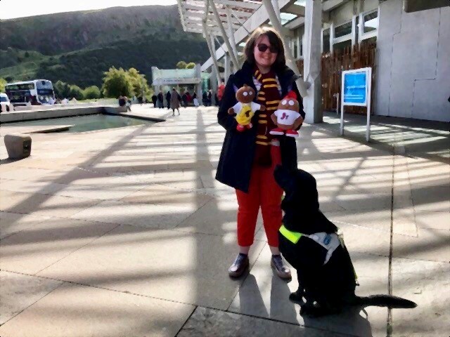 image description: Eilidh standing in front of the Scottish Parliament with her guide dog who is a black lab/retriever. She is holding the mascots for Haggeye Jnr and Haggeye Tots