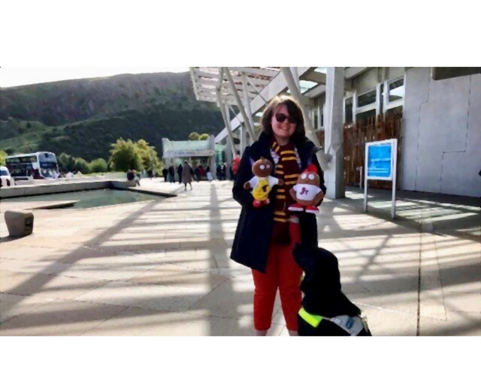 image description: Eilidh standing in front of the Scottish Parliament with her guide dog who is a black lab/retriever. She is holding the mascots for Haggeye Jnr and Haggeye Tots