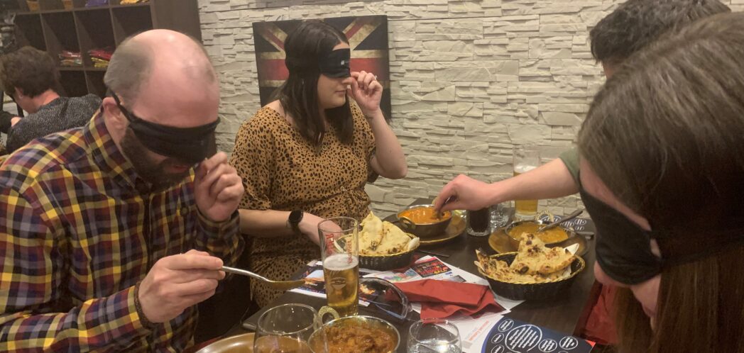 Man in plaid shirt and woman in yellow dress sit facing another man and woman. They are all wearing blindfolds while they eat indian food