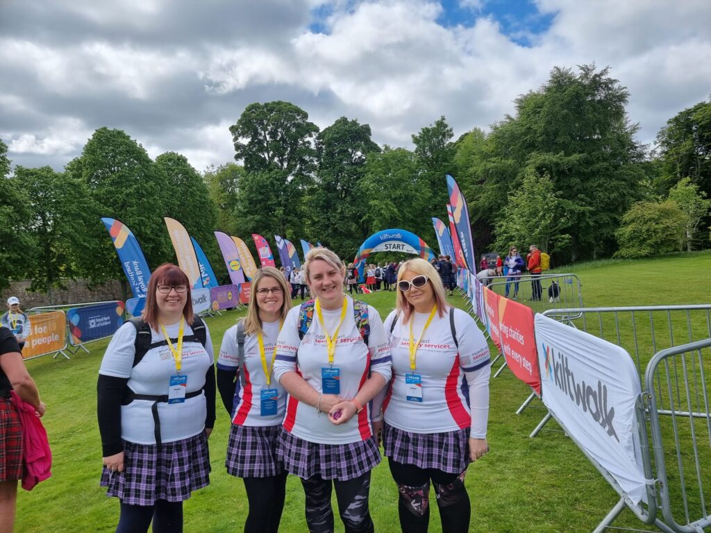 Lauren Paterson and the other three members of WhattheHillBanff team, Laurieann, Dianne and kim stand in the lead up to the start line for the kiltwalk, surrounded by a number of banners. they are all wearing NESS t-shirts, the kiltwalk pass, and purple tartan skirts. 