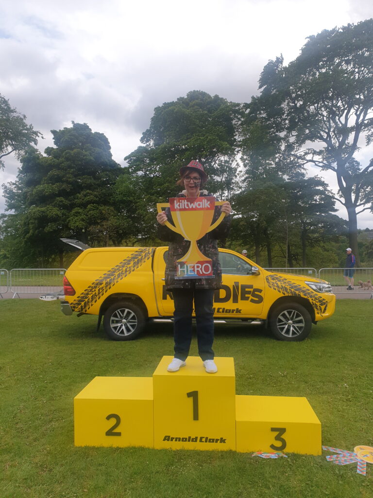 Nicola Young stands on the yellow winners podium in the number one position, holding a cut out trophy that says kiltwalk hero. She is wearing a tartan hat. 