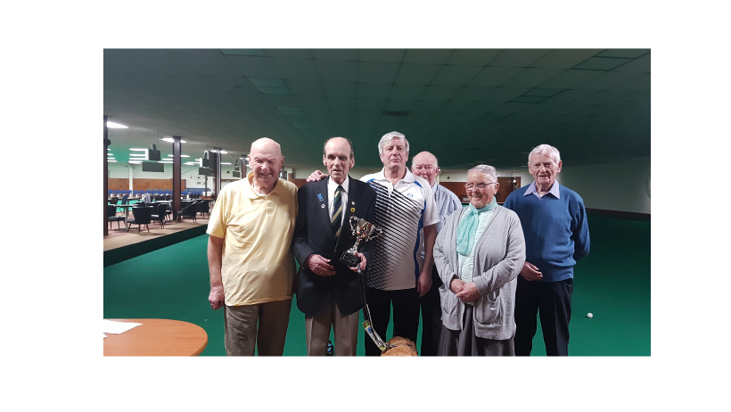 group of 6 older adults stand togehter. the man second from the left wears a shirt, tie and blazer and carries a trophy