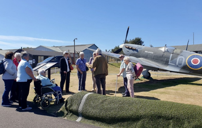 Ten group members stand outside in glorious weather next to an old fighter plane hearing about its history
