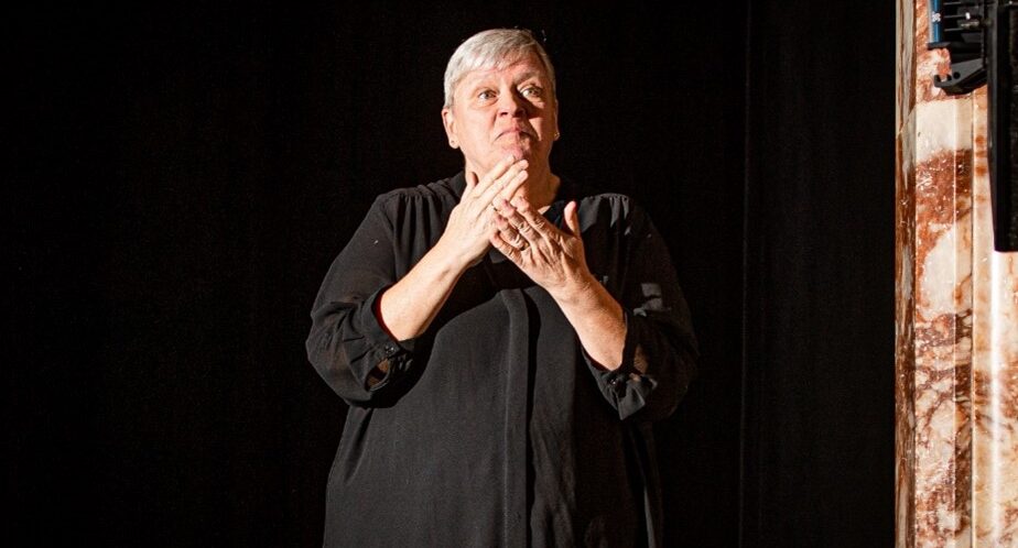 Lelsey Crerar, dressed in black against a black background, interprets at the theatre