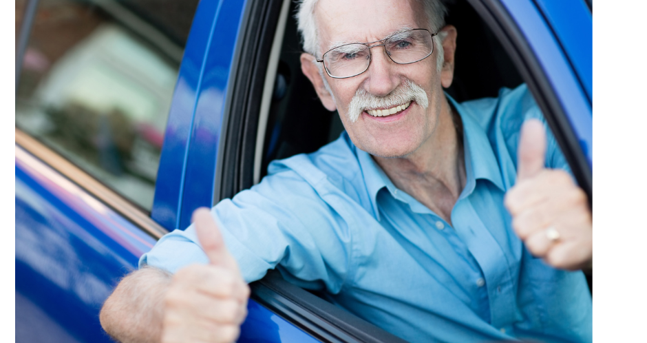 Older man leans out of car window smiling and holding both thumbs up
