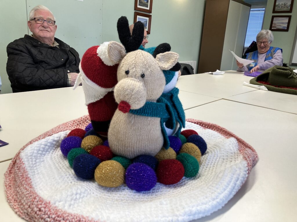 Close up of knitted Christmas scene, with Rudolph the Red-nosed Reindeer in the foreground, Father Christmas can be seen side on to the left and another reindeer can just be made out behind them.