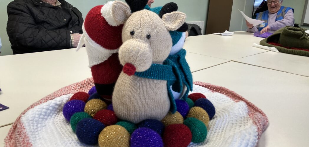 Close up of knitted Christmas scene, with Rudolph the Red-nosed Reindeer in the foreground, Father Christmas can be seen side on to the left and another reindeer can just be made out behind them.