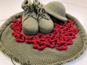 Close up of knitted remembrance day item, with khaki green knitted base, a surround of red poppies, khaki knitted boots and helmet 