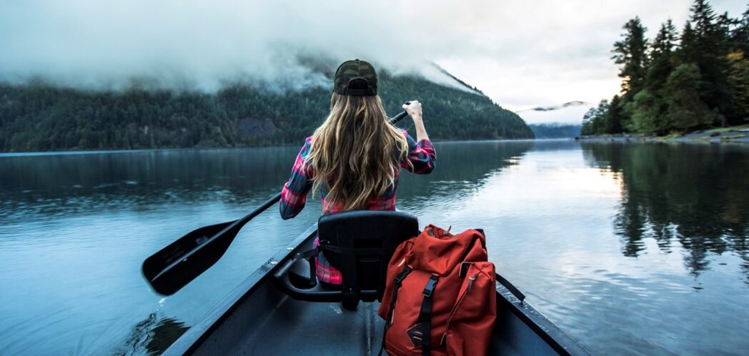 Young woman seen from the back paddles in a canoe on a beatiful lake surrounded by forested hills and low lying mist. A red back pack is seen in the canoe.