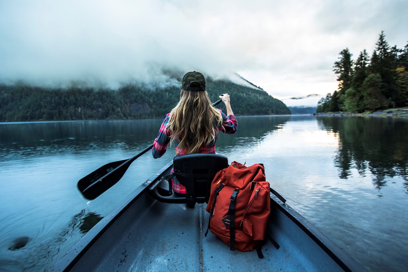 Young woman seen from the back paddles in a canoe on a beatiful lake surrounded by forested hills and low lying mist. A red back pack is seen in the canoe.