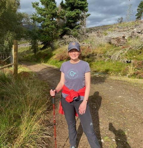 Valerie Goggin wearing hiking gear, baseball cap and dark glasses stands holding a walking cane against a backdrop of tree covered hills