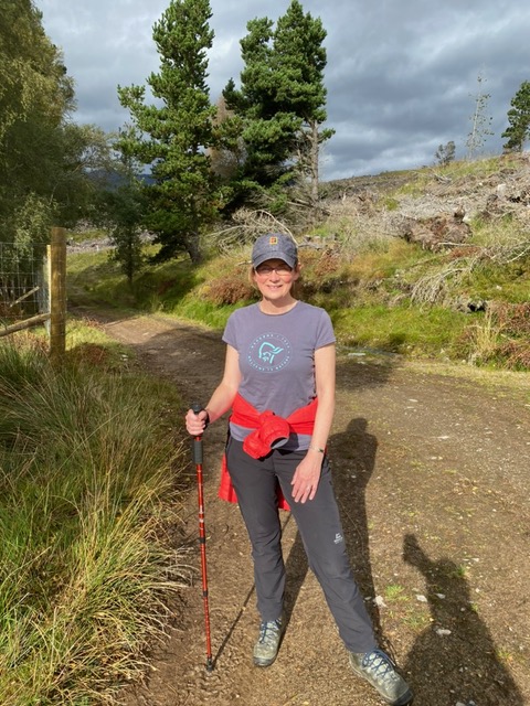 Valerie Goggin wearing hiking gear, baseball cap and dark glasses stands holding a walking cane against a backdrop of tree covered hills