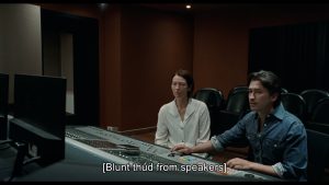 A young man and a young woman sit in from of a large mixing deck, with descriptive captions in square brackets that say "blunt thud from speakers".