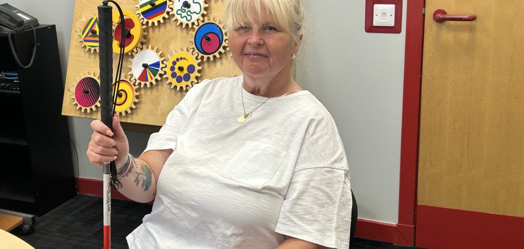 Susan smiles holding her red and white cane. In the background, is a wall mounted interactive sensory board.
