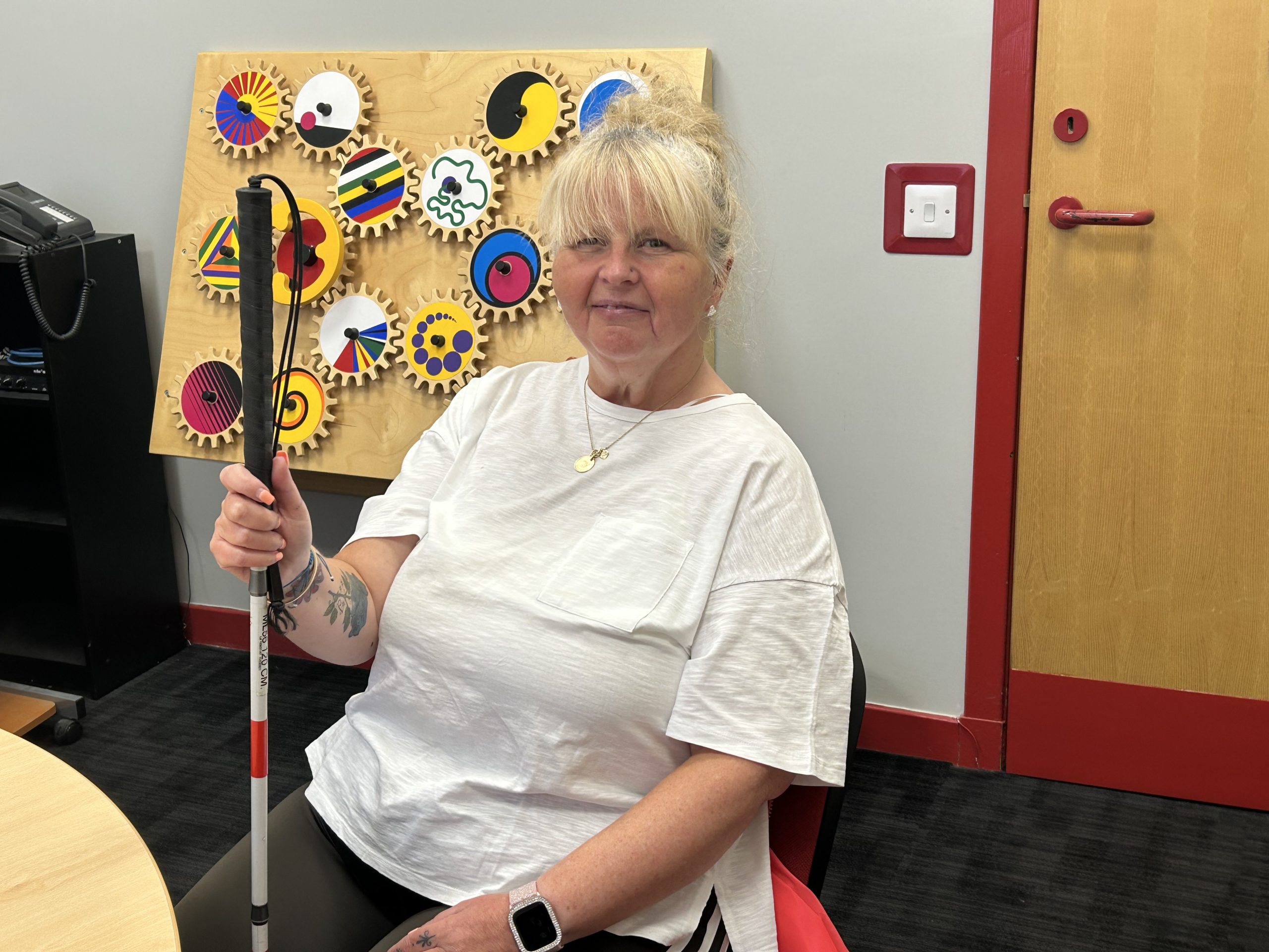 Susan smiles holding her red and white cane. In the background, is a wall mounted interactive sensory board.