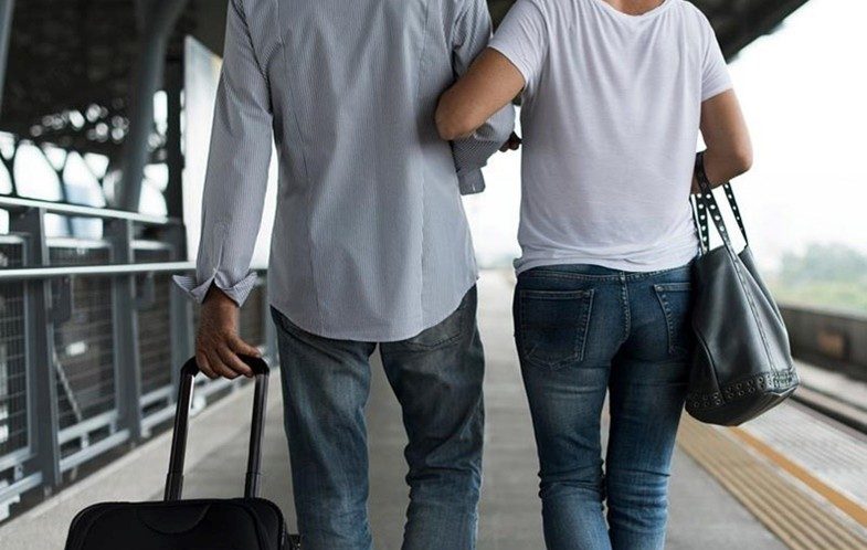 Young woman links arms with a young man as they walk away from us on a train platform