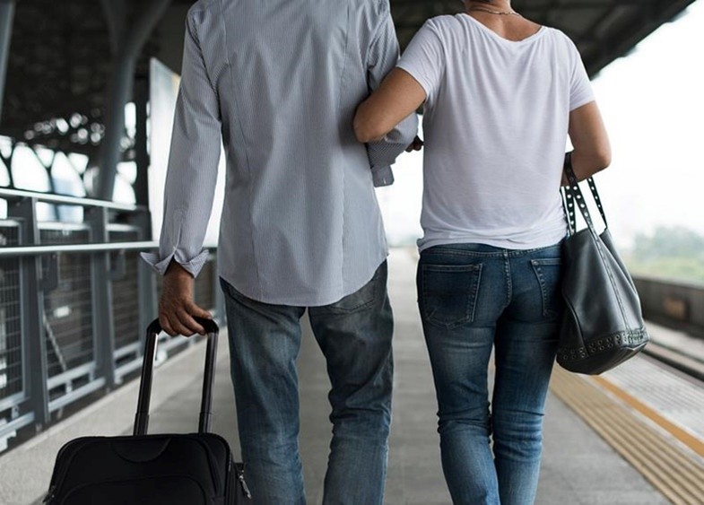 Young woman links arms with a young man as they walk away from us on a train platform