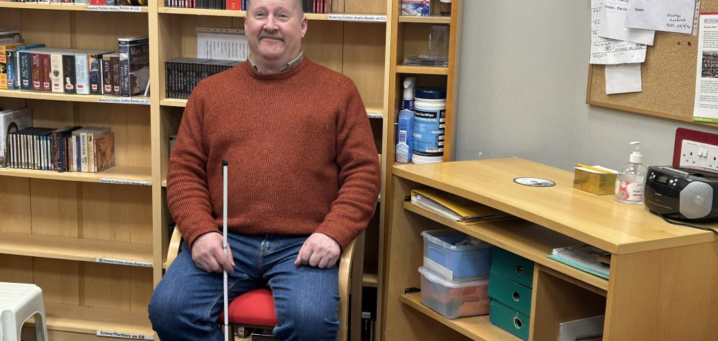 Kevin Shaw sits on a chair, holding his symbol cane in his right hand. A bookcase is behind him and a desk is to his left.