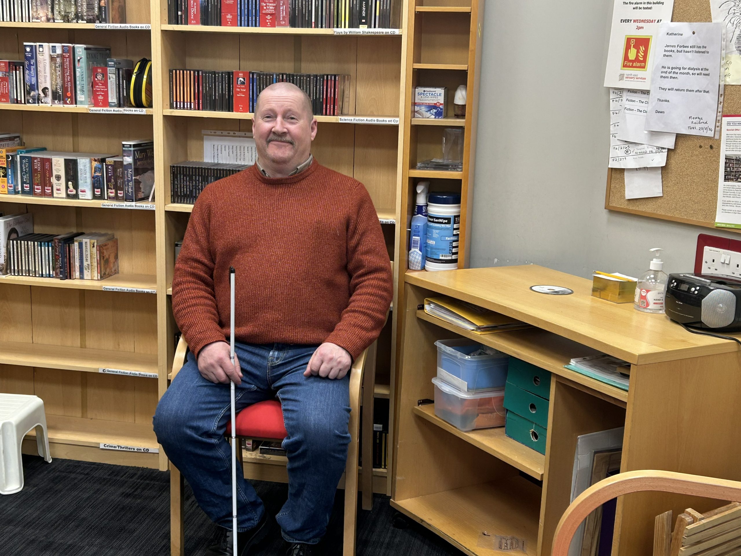 Kevin Shaw sits on a chair, holding his symbol cane in his right hand. A bookcase is behind him and a desk is to his left.