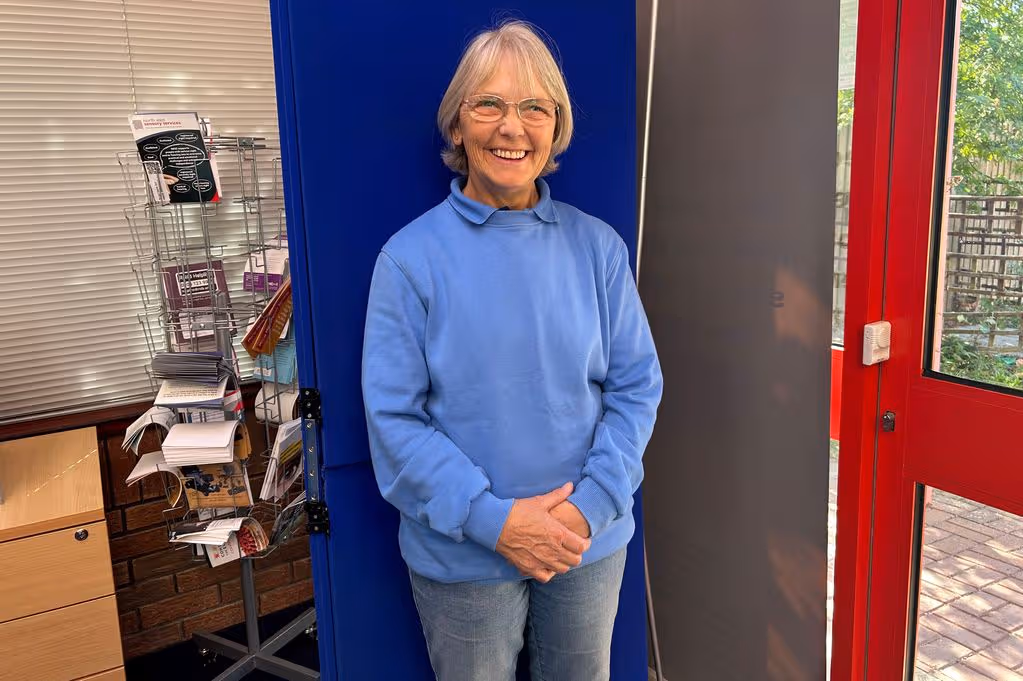 Ethna Taylor stands smiling in front a blue background with a leaflet holder behind her on her right.